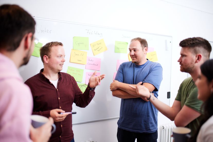 Ein Team steht vor einem Whiteboard mit vielen bunten und gruppierten Post its und diskutiert.