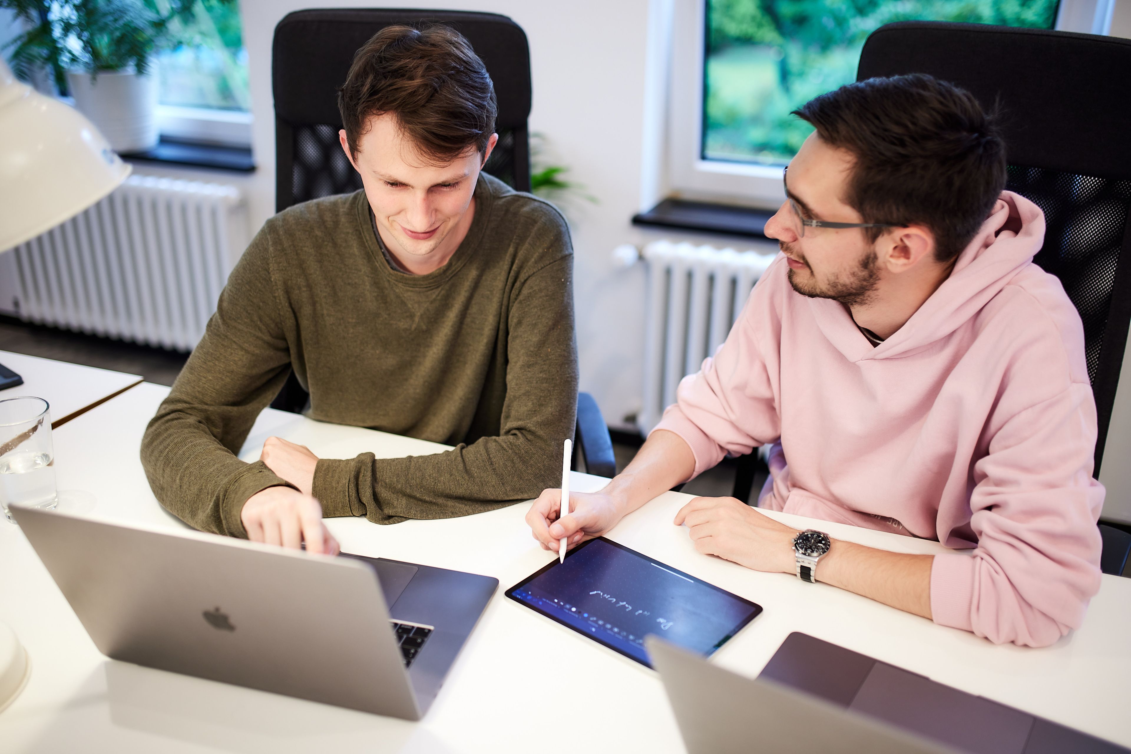 Zwei Kollegen sitzen an einem Bürotisch nebeneinander und besprechen sich vor einem Laptop und an einem Tablet.